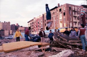 Bronx kids play and do flips on debris in a vacant lot, surrounded by burned-out buildings.